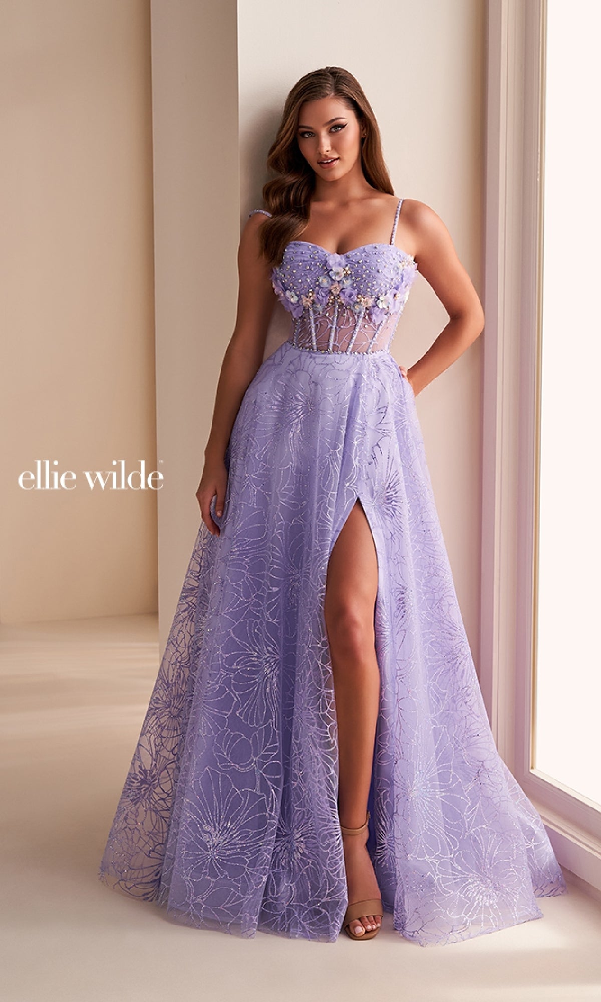 A woman stands by a window wearing the Ellie Wilde EW36218 long prom dress, an A-line lavender gown with floral lace, 3D flowers, a sheer corset bodice, spaghetti straps, and a thigh-high slit.